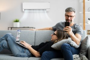 A man and a woman sitting on the couch. The woman has her head in the man's lap and is using a laptop. The man is looking at his phone. There is an air conditioner running behind them