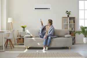 Woman is sitting on a couch and is using a remote control to turn on the AC unit behind her.