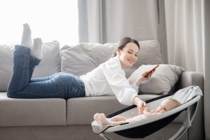 Young mother lying on a sofa next to infant in bouncy seat